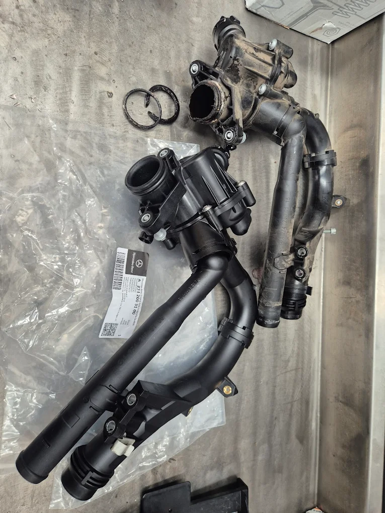 Mercedes-Benz vehicle parts including a black coolant pipe assembly and a used component, on a workshop surface