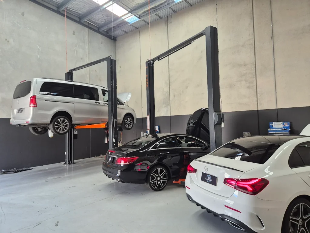 Mercedes-Benz vehicles in a modern garage setting, featuring a silver van on a lift, a black car with its hood open, and
