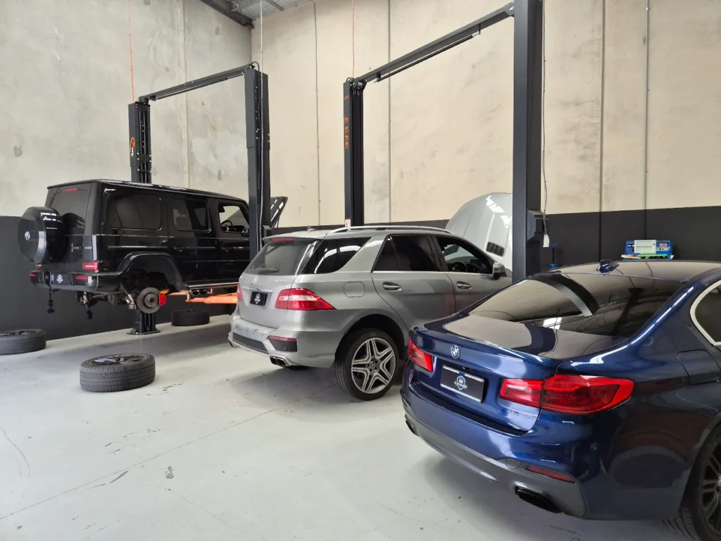 Mercedes-Benz vehicles in a modern garage, featuring a black G-Class SUV on a lift, a silver ML-Class SUV, and a blue BM