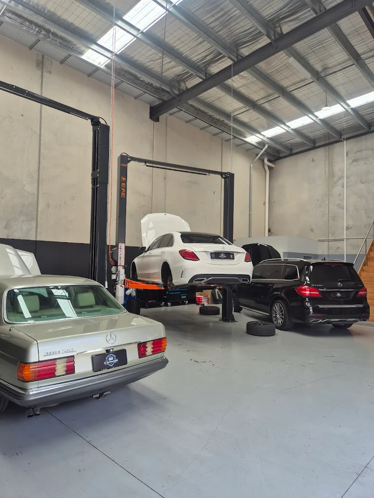 Mercedes-Benz vehicles in a garage, including a white model on a lift for maintenance, with additional black and silver
