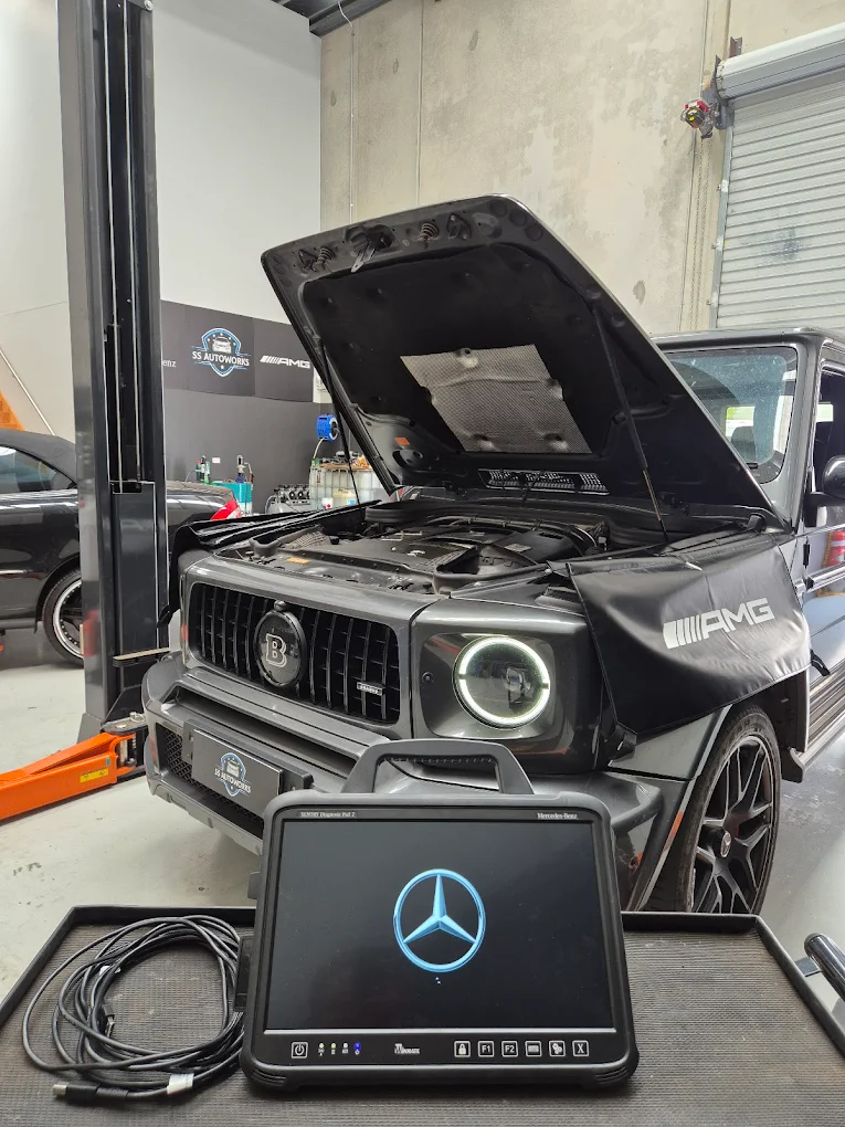 Mercedes-Benz vehicle in garage with open hood, diagnostic tablet displaying logo, tools and equipment visible