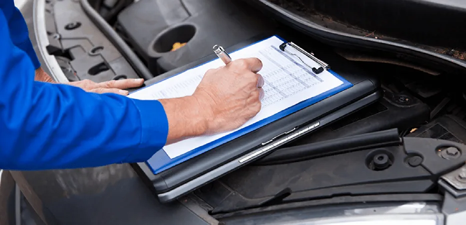 Mechanic in blue uniform writing on a clipboard while inspecting a vehicle's engine